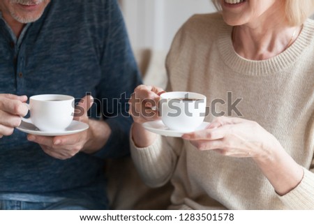 Positive middle aged spouses sitting at home holding cups with black tea beverage. Mature senior wife and husband spending time together drinking afternoon coffee and chatting, close up cropped image