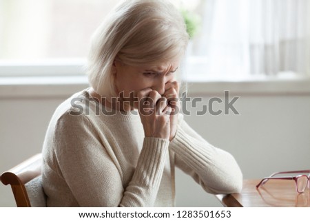 Lonely aged blonde woman sitting at table at home with sad face expression holding hands near face feels unhappy miserable and desperate. Physical and psychological mental health problems of elderly Royalty-Free Stock Photo #1283501653