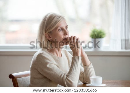 Woman spend time at home alone sitting at table with cup of tea folds hands on chin lost in thoughts. Old lonely female has health problem or thinking about life, reminiscing the past relive memories Royalty-Free Stock Photo #1283501665