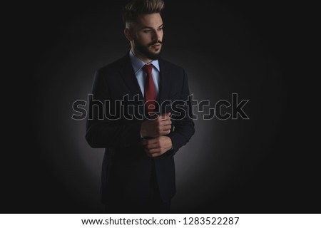 portrait of surprised businessman arranging suit cuffs and looking down to side while standing on black background