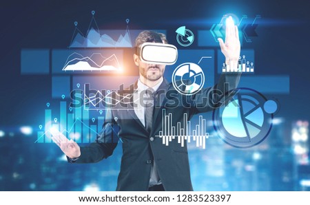 Businessman with beard wearing dark suit and vr glasses interacting with business infographics interface over blurred background. Toned image double exposure