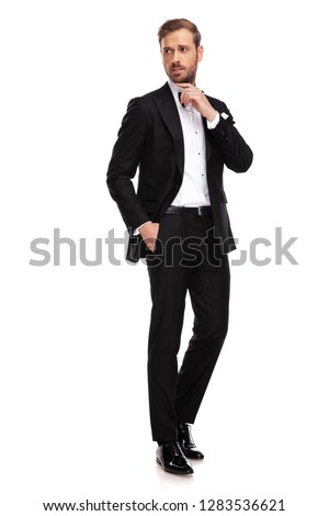 relaxed businessman in black suit standing on white background with hand in pockets looks and points to side, full length picture
