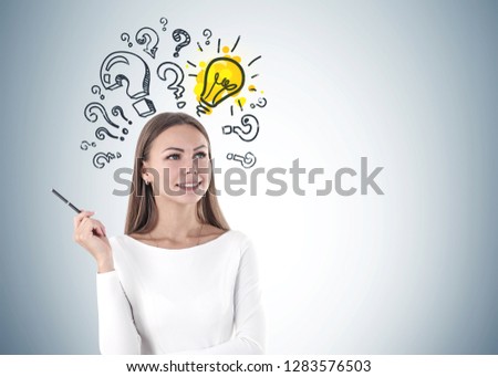 Smiling young woman with fair hair wearing white and holding a pen standing near gray wall with question marks and lightbulb on it. Mock up