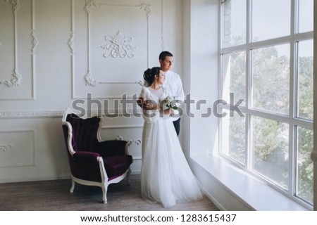 A beautiful bride and a young groom are standing near a window in a white studio. Wedding in the interior. Wedding portrait of newlyweds in love. The photo.