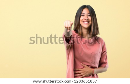 Young girl with pink shirt pointing with finger at someone and laughing a lot on yellow background