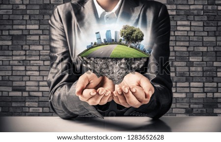 Businessman in suit keeping green island with skycraper city in hands with gray brick wall on background.