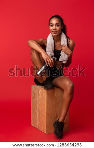 Portrait of athletic african american woman in black sportswear sitting on box with towel and water bottle isolated over red background