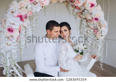 A beautiful bride and a young groom are sitting in a white studio on a swing. Wedding in the interior. Wedding portrait of newlyweds in love. The photo.