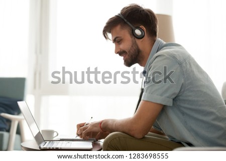 Happy man wearing headset study online learning foreign english language or watch webinar make notes, smiling male student in headphones looking at laptop elearning in internet write down information Royalty-Free Stock Photo #1283698555