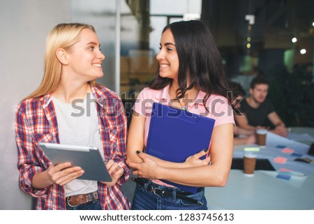 Picture of young blonde and brunette models stand together and smile. They look happy. Young women hold electronic and plastic tablets in hands. Two young men work behind them.