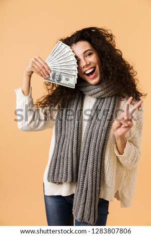 Cheerful young woman wearing winter scarf standing isolated over beige background, holding money banknotes