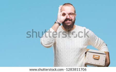 Young hipster man wearing glasses, moving holding moving box over isolated background with happy face smiling doing ok sign with hand on eye looking through fingers