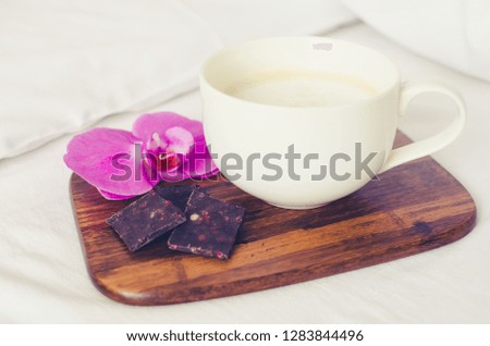 Cup of coffee cappuccino with chocolate and flower on wooden board on a bed on cozy lazy sunday. White bedding sheet, blanket and pillows. Good morning concept. Enjoy slow life.