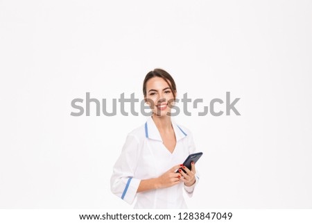 Confident young woman doctor wearing uniform standing isolated over white background, using mobile phone