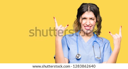 Young adult doctor woman wearing medical uniform shouting with crazy expression doing rock symbol with hands up. Music star. Heavy concept.