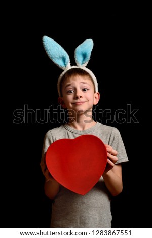 Funny blond boy in hare ears holds heart shape red box on black background. Love and family concept. Vertical frame.