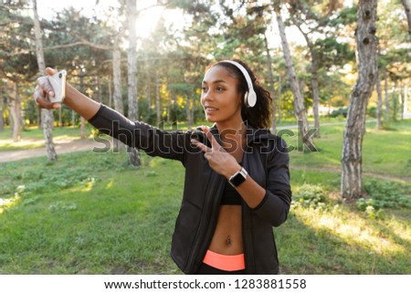 Portrait of happy woman 20s wearing black tracksuit and headphones taking selfie photo on cell phone while walking through green park