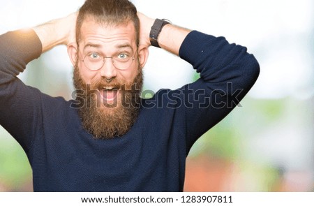 Young blond man wearing glasses and turtleneck sweater Crazy and scared with hands on head, afraid and surprised of shock with open mouth