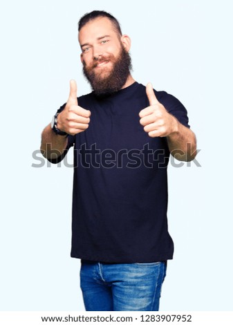 Young blond man wearing casual t-shirt approving doing positive gesture with hand, thumbs up smiling and happy for success. Looking at the camera, winner gesture.