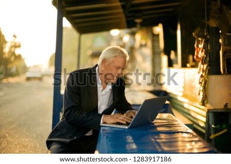 Businessman types on a laptop as he sits at a table outside a cafe on a rural road.