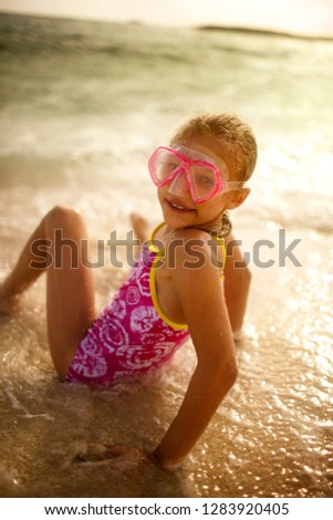 Portrait of a smiling young girl wearing swimming goggles while sitting in shallow water at the beach.
