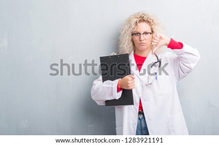 Young blonde doctor woman over grunge grey wall holding clipboard with angry face, negative sign showing dislike with thumbs down, rejection concept