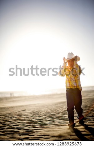 Young boy wearing a cowboy hat while walking along a sandy beach in the sun.