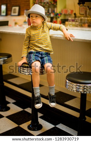 Young boy wearing a porkpie hat perches on a tall stool at a restaurant counter.