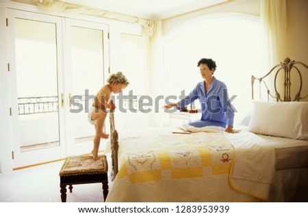 Side view of a kid climbing onto the bed as his mother looks on.