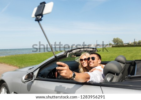 road trip, leisure, technology and people concept - happy couple in convertible car taking picture by smartphone on selfie stick