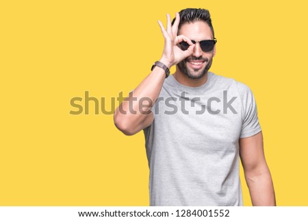 Young handsome man wearing sunglasses over isolated background with happy face smiling doing ok sign with hand on eye looking through fingers