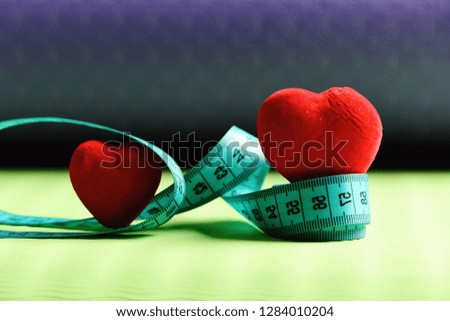 Workout and sport concept. Shaping and fitness. Heart decorations and flexible ruler lying on yoga mat. Measuring tape roll in cyan color and soft red hearts on green and purple background, defocused