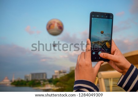 A woman is taking a picture of a hot air balloon flying into the sky using a mobile phone