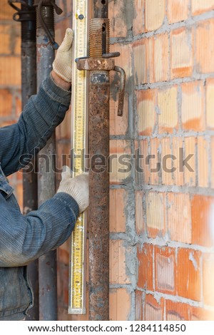 Formwork construction concept. Cropped and vertical photo of mature specialist measuring pipe height in unfinished house using special ruler