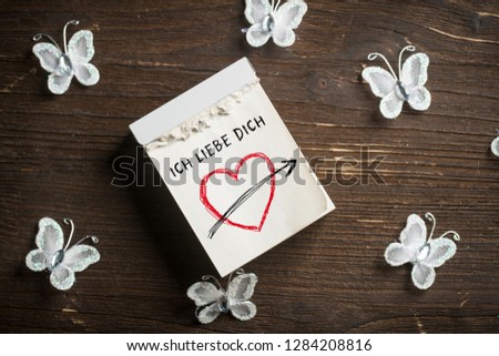 message 'I love you' (in German) on a tear-off calendar surrounded by butterflies on wooden background