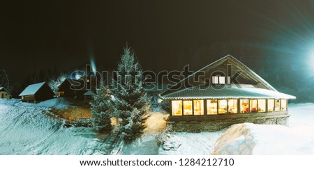 Photo of a cozy wooden house covered in snow near mountains with the lights turn on