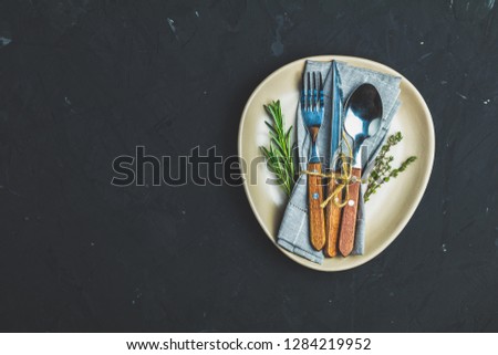 Rustic vintage set of cutlery knife, fork and spoon in light ceramic plate. Black stone concrete surface background. Top view, copy space.
