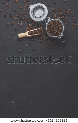 Glass jar with roasted beans and wooden scoop on black stone concrete textured surface background. Top view with copy space for your text.