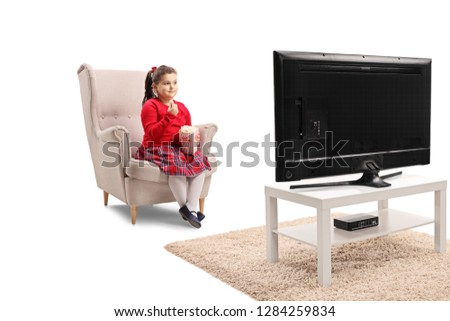 Full length shot of a little girl sitting in an armchair with popcorn and watching TV isolated on white background