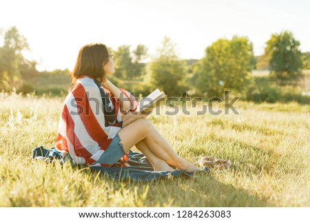 Mature woman reads a book, rests on the nature. On the shoulders American flag, background sunset, rustic landscapes, green meadow.