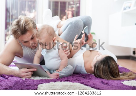 Happy parents laying down on the floor and playing with their baby boy. Computer era. Father shows cartoons to his child. Mother playing on the mobile phone.