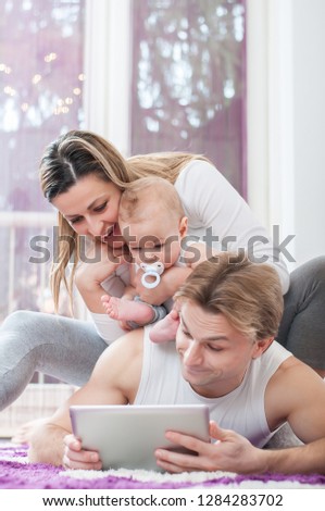 Happy parents laying down on the floor and playing on tablet with their baby boy. Father shows a cartoons to his child.