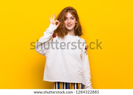 Young redhead woman over yellow wall showing an ok sign with fingers