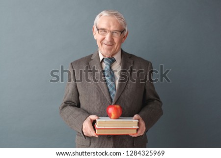 Senior male teacher wearing eyeglasses studio standing isolated on gray wall holding books and apple looking camera smiling friendly
