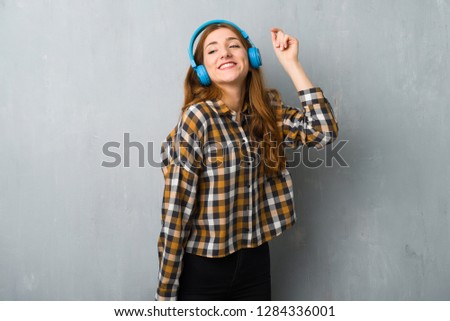 Young redhead girl over grunge wall listening to music with headphones and dancing