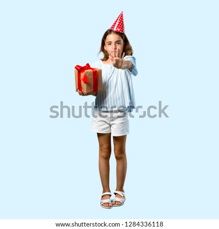 Full body of Little girl at a birthday party holding a gift making stop gesture with her hand on blue background
