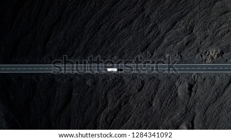 The road with white lines is surrounded by black volcanic lava. Sticking rocks. Dangerous adventure of incredible beauty. Top view, drone footage. Minimalistic landscape. White car in motion. Royalty-Free Stock Photo #1284341092