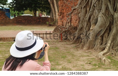 Female Tourist Taking Pictures of Buddha Image's Head Trapped in Bodhi Tree Roots, Wat Mahathat Ancient Temple, Ayutthaya Historical Park, Archaeological site in Thailand