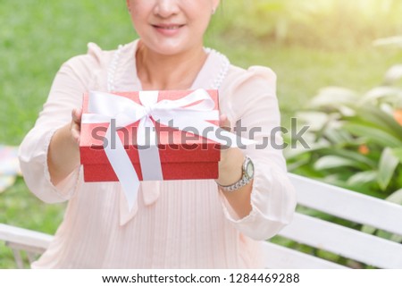 Senior Asian woman showing red present box with white ribbon outdoor.