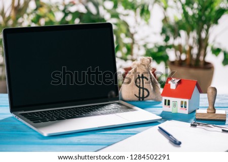 selective focus of pen, clipboard, laptop with blank screen near stamp, house model and moneybag with dollar sign on wooden desk, mortgage concept